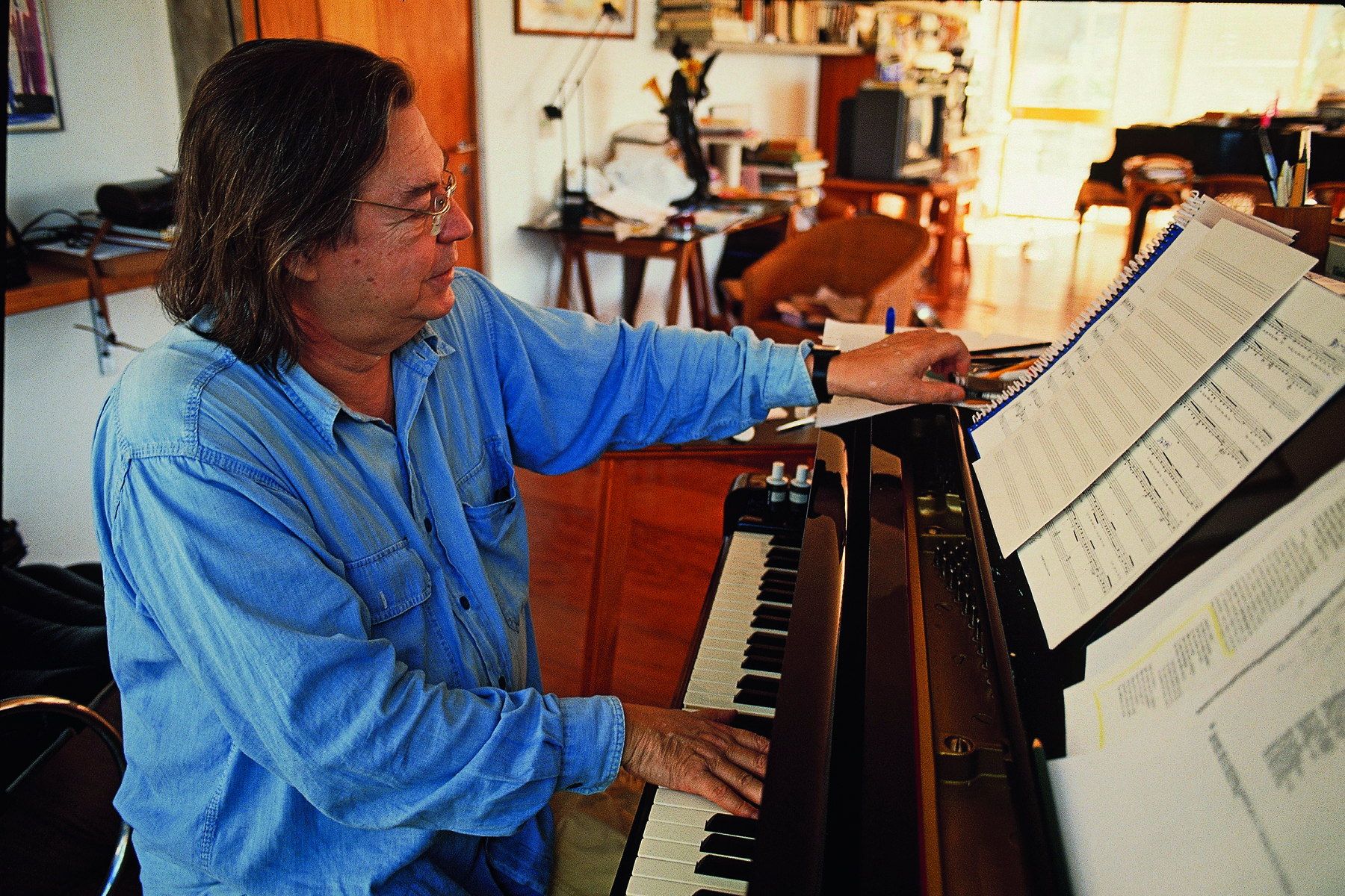 Fotografia. Homem de cabelo médio liso castanho e óculos de armação arredondada, vestindo camisa azul. Ele está de perfil, tocando piano enquanto observa uma partitura disposta na parte de cima do piano.