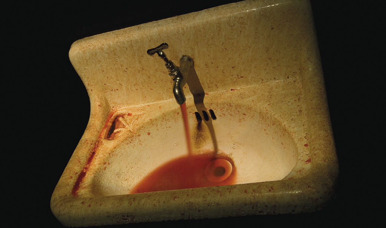 Fotografia. Instalação que apresenta uma pia suja com a torneira aberta de onde escorre um líquido vermelho.