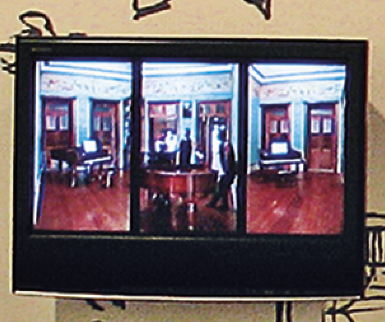 Fotografia. Detalhe da videoinstalação com monitores na parede que tem um desenho de cômodos de uma casa pintados. Detalhe de um dos monitores, que mostra três imagens de diferentes ângulos de uma sala com paredes azuis, piso de madeira e duas portas. Há um piano próximo às portas.