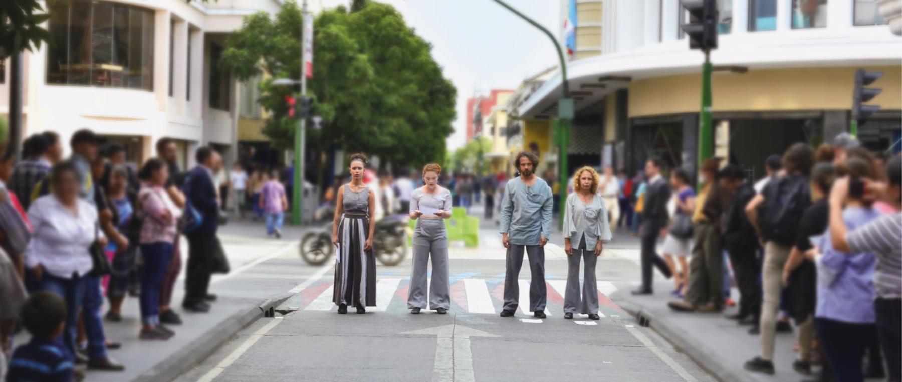 Fotografia. Quatro artistas se apresentam em uma via pública. Eles estão em cima de uma faixa de pedestres, perfilados. Todos usam roupas com tons de cinza. Há pessoas nas calçadas observando a cena.