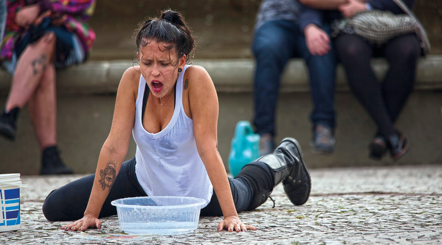 Fotografia. Artista faz performance na rua. Ela está de camiseta branca e calça preta e está em frente a uma bacia com água, apoiada com as mãos no chão. Seu rosto e camiseta estão molhados.