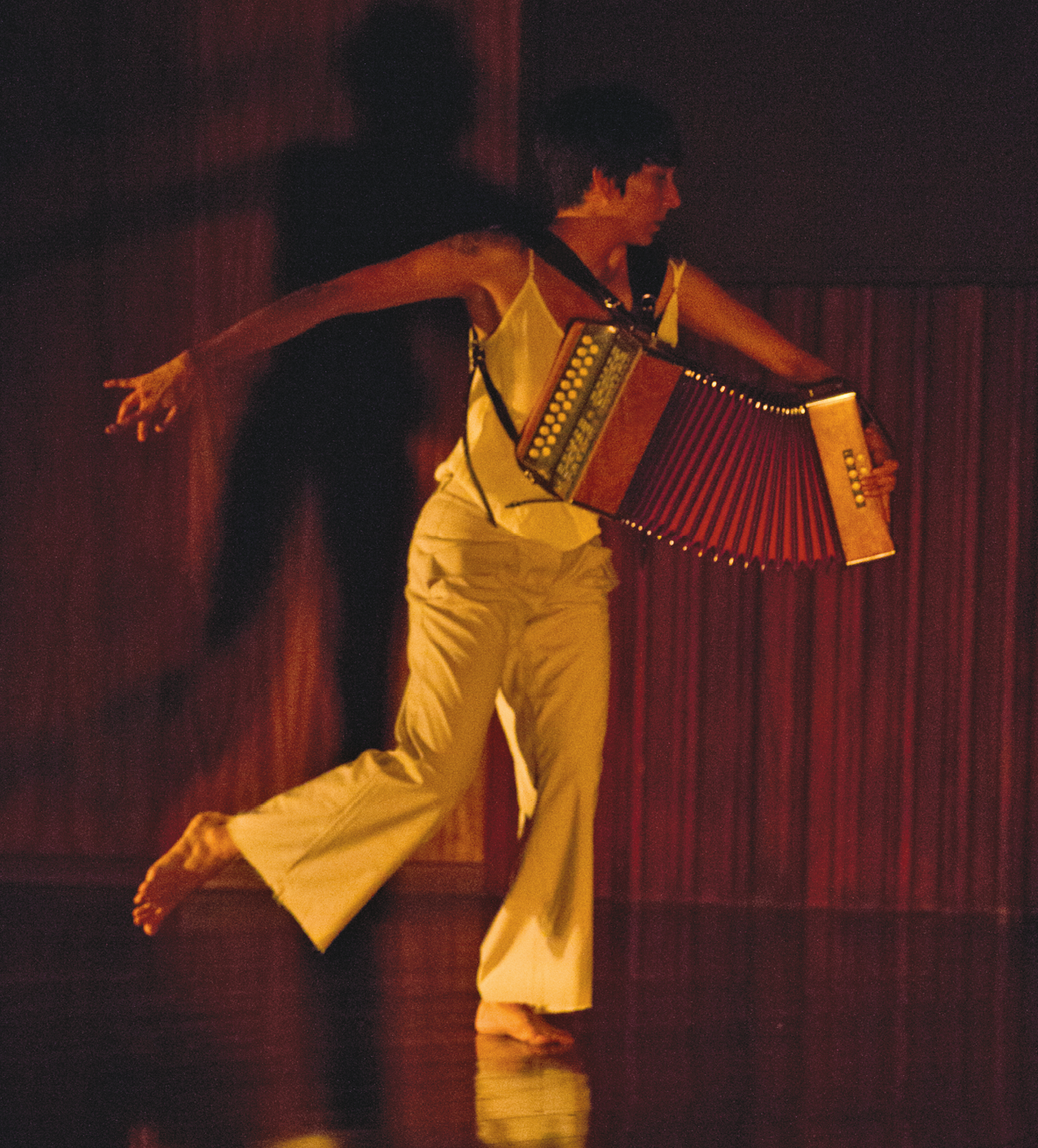 Fotografia. Mulher em um palco, vestida com roupas brancas, carrega acordeão e realiza movimento de dança, com uma das pernas suspensa e um braço levantado.