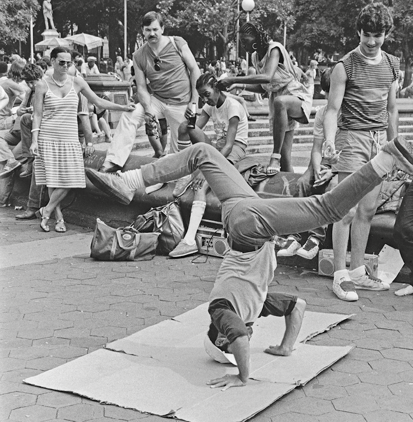 Fotografia em preto e branco. Homem dança break em uma praça. Ele está vestido com camiseta branca e calça jeans, com a cabeça e as mãos apoiadas no chão, e as pernas abertas ao alto. Ao redor, pessoas observam a apresentação.