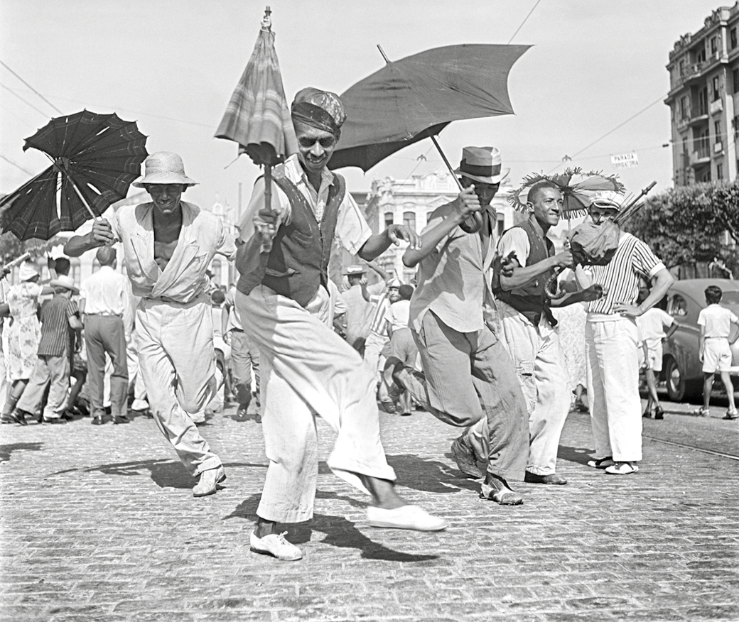 Fotografia em preto e branco. Grupo de pessoas dança frevo com guarda-chuvas. Os homens usam camisa e calças sociais, com chapéus, em uma rua de paralelepípedo.