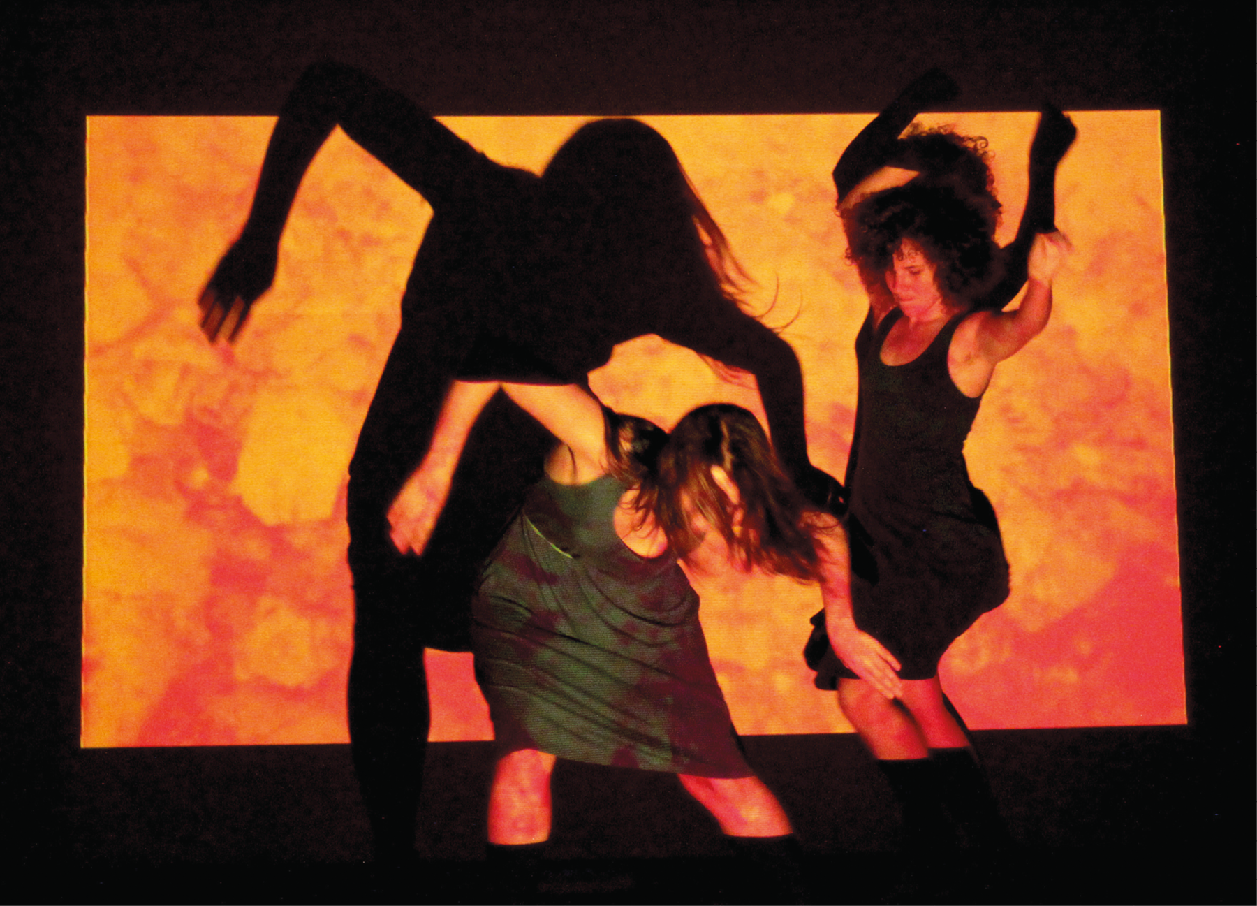 Fotografia. Duas mulheres dançam em frente a um telão no qual é projetado um desenho abstrato em tons de laranja. Elas vestem vestidos escuros e estão em movimento. Com a luz, suas sombras são projetadas no telão.