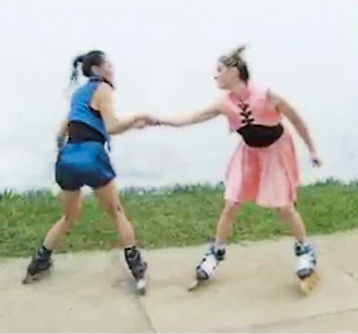 Fotografia. Duas adolescentes (à esquerda, a menina veste camiseta azul e shorts; à direita, a menina usa vestido em tons de rosa) estão de mãos dadas, uma de frente para a outra, no centro de uma calçada, andando de patins. Ao fundo, o mar.