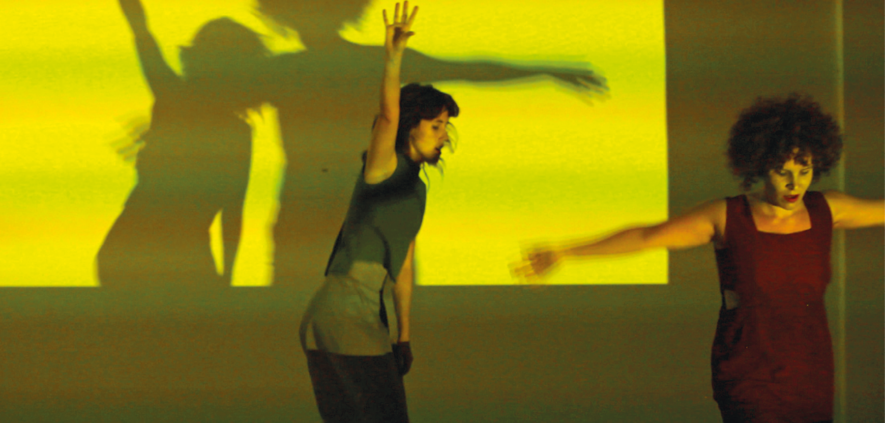 Fotografia. Duas mulheres dançam em frente a um telão no qual é projetado uma luz em tons de verde-claro e amarelo. Elas vestem vestidos escuros e estão em movimento, com os braços abertos. Com a luz, suas sombras são projetadas no telão.