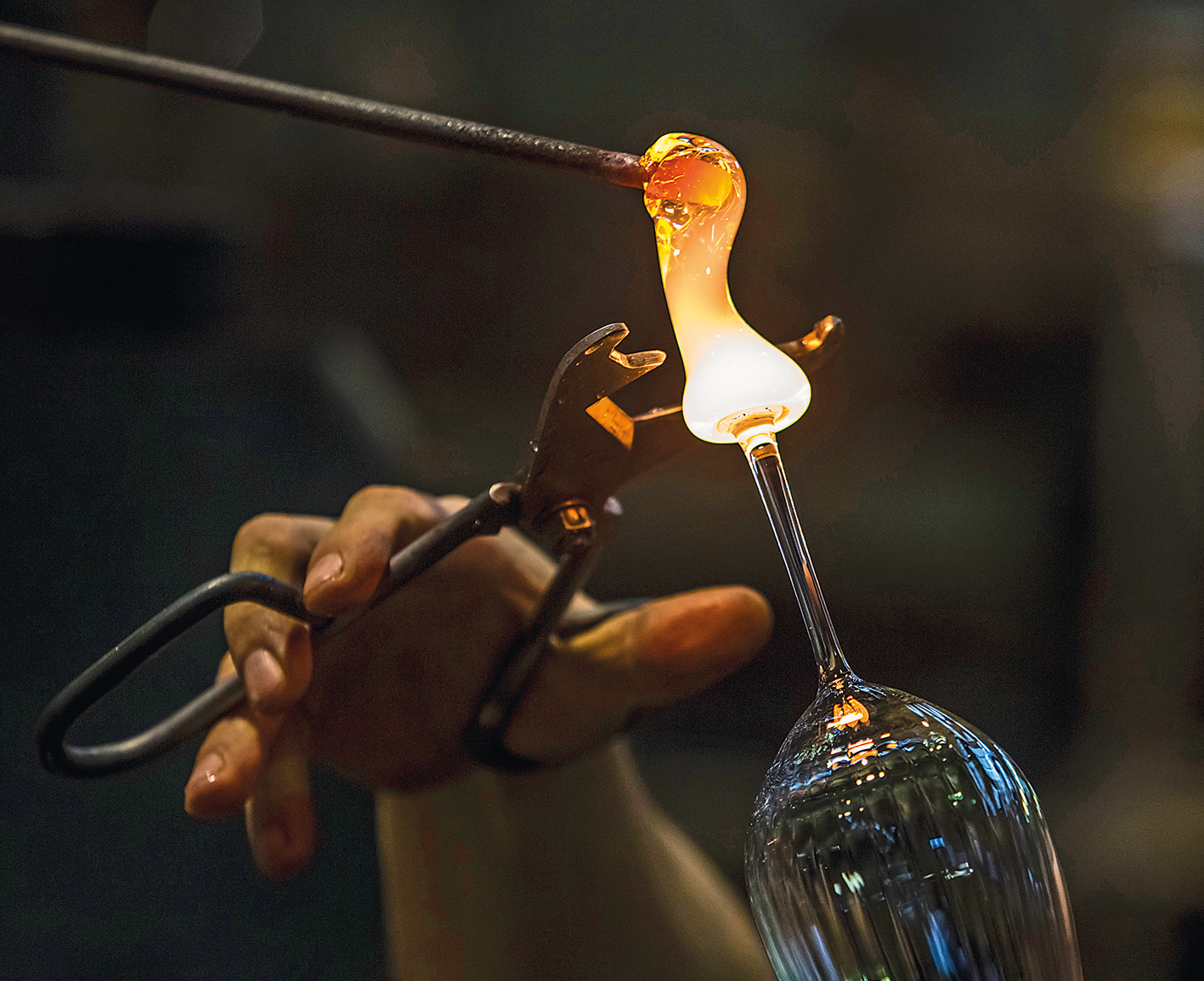 Fotografia. Mão de uma pessoa segurando uma ferramenta, semelhante a uma tesoura, na direção da parte inferior de uma taça de vidro que ainda está sendo moldada, maleável e quente, com cor amarelada.