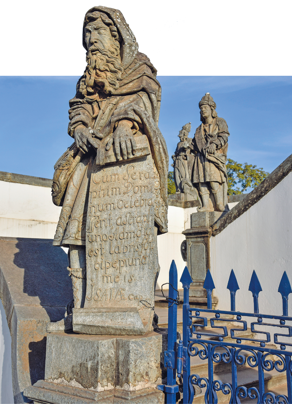 Fotografia. À frente de uma construção, próximo a um portão, uma estátua feita de rocha. Ela representa um homem que usa capuz, tecidos envoltos no corpo, cabelo e barba longos. Segura um papiro com inscrições. Ao fundo, outras duas estátuas que representam pessoas em pé.