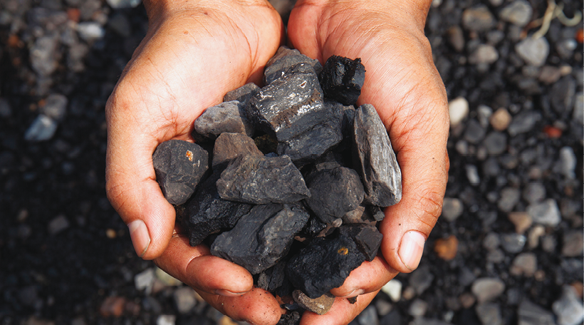 Fotografia de duas mãos juntas segurando pequenas pedras pretas e irregulares. Ao fundo, outras pedras pretas.