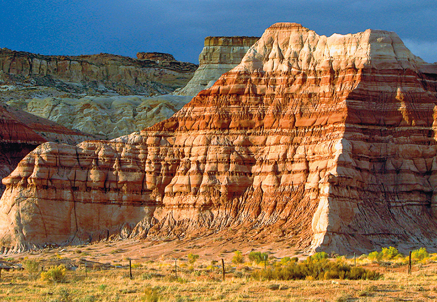 Fotografia de uma formação rochosa com muitas camadas de cores amarronzadas, mais claras e mais escuras. Ao fundo, mais rochas. O céu está azul.