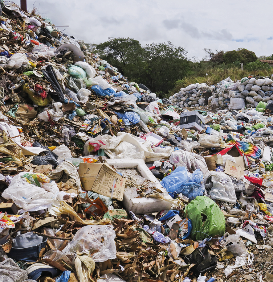 Fotografia de uma área a céu aberto com muitos resíduos acumulados: sacos plásticos, papelão, objetos descartados, folhas, dentre outros.