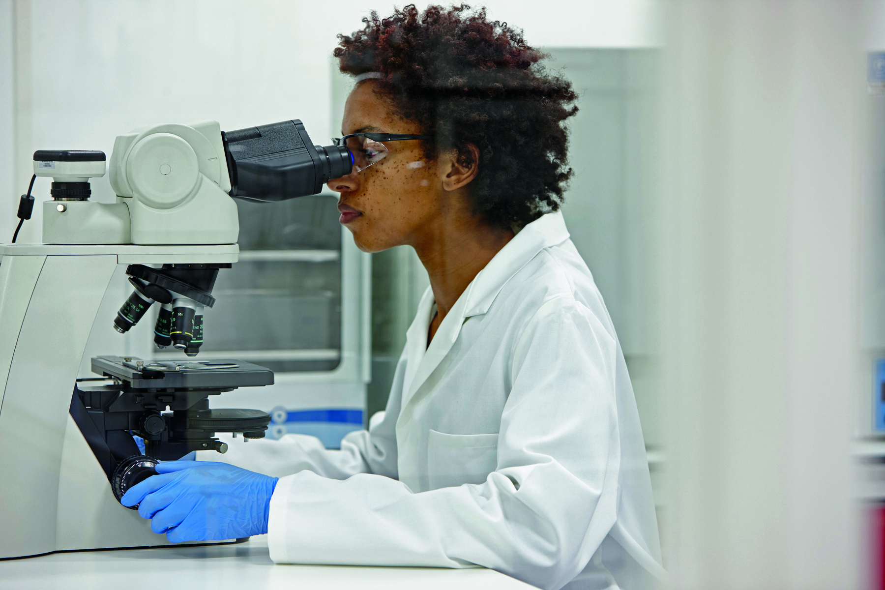 Fotografia. Uma mulher negra sentada, usando jaleco branco, luvas azuis e óculos. Ela está olhando por um microscópio.