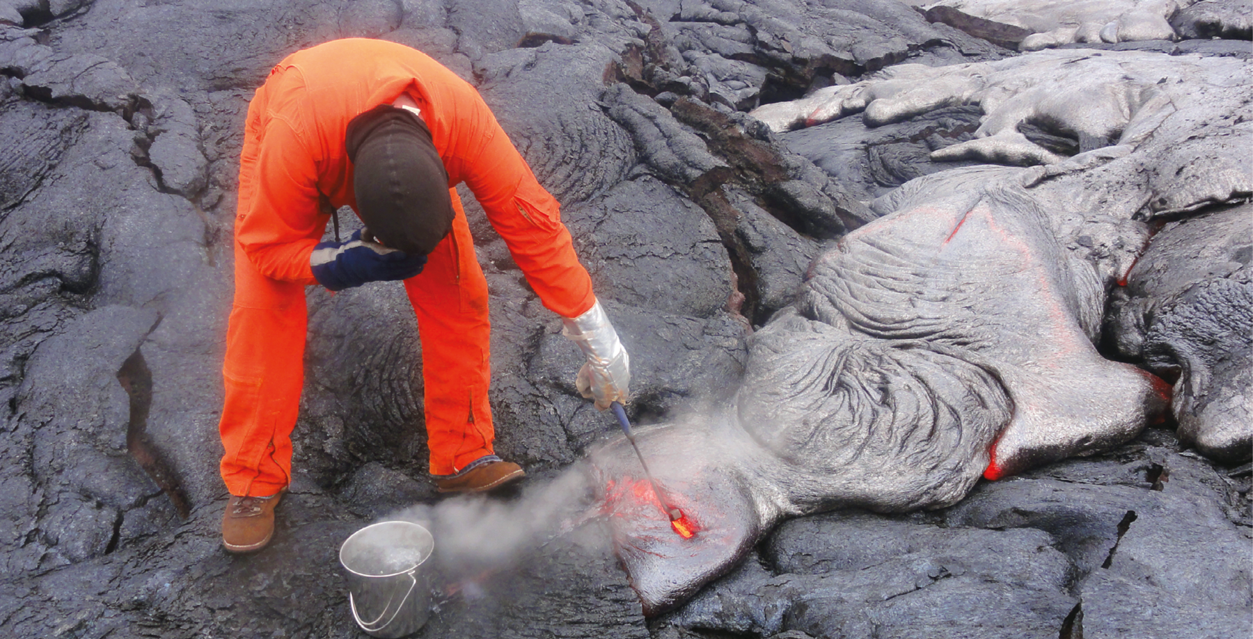 Fotografia. Um homem, usando roupa toda laranja, touca, sapatos e luvas, está agachado, segurando um equipamento sobre lava que está no chão. Ao lado, um balde. Ao redor, o chão é escuro e irregular.