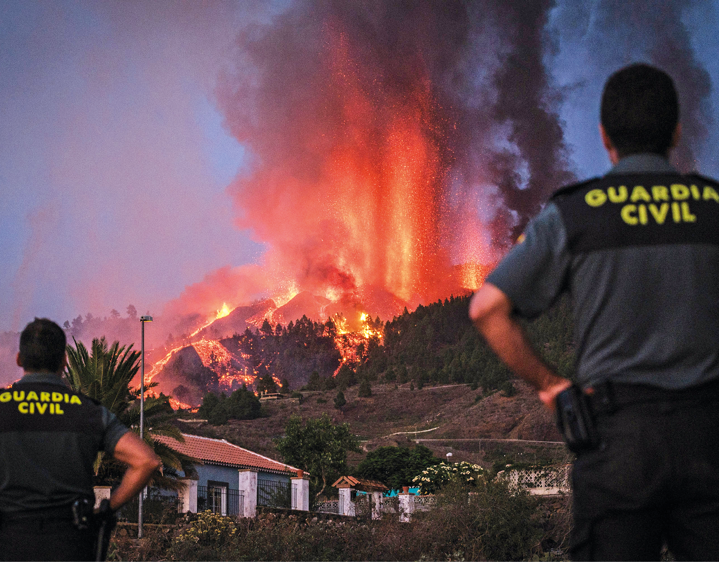 Fotografia. Dois policiais brancos em pé, de costas, olhando para o fundo, na direção de um vulcão que está em erupção, expelindo lava. Ao redor, uma área com casas, montanhas e vegetação.