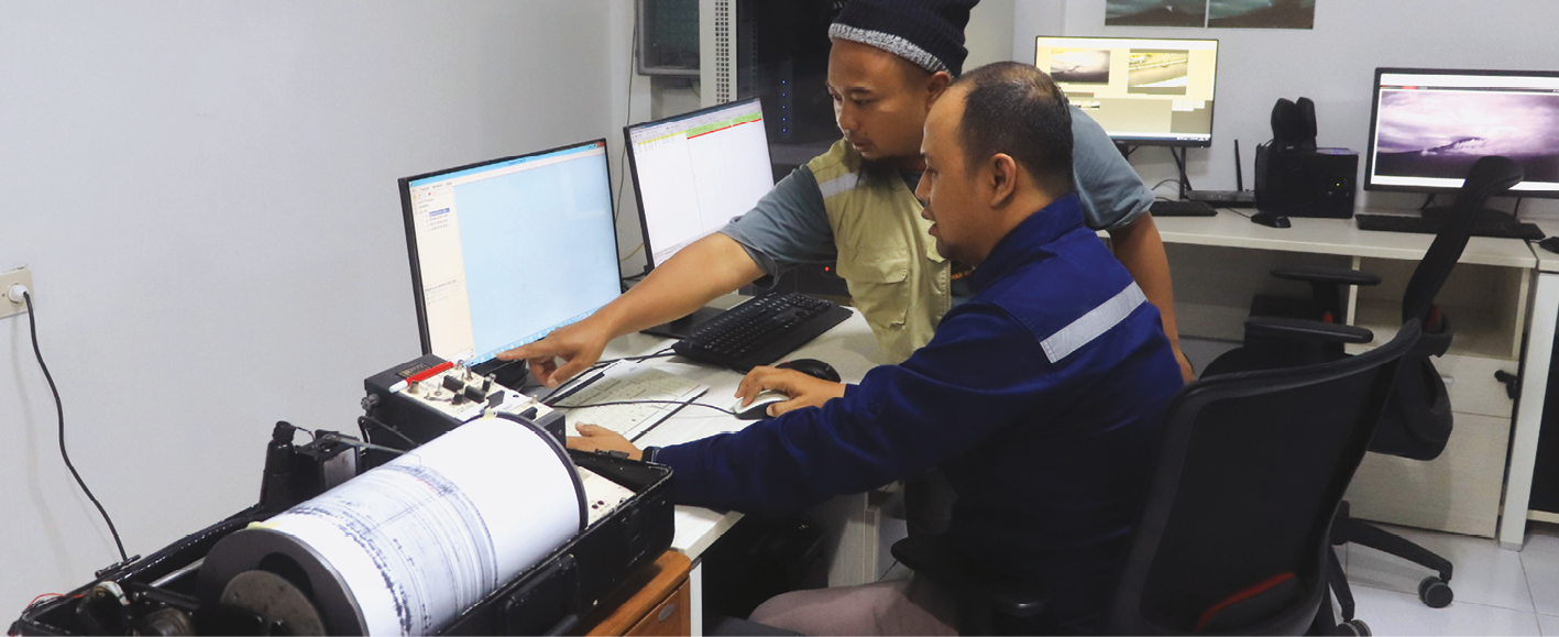 Fotografia. Um homem asiático sentado em uma cadeira, em frente a um computador, manuseando-o. Ao lado, um sismógrafo em funcionamento, imprimindo relatório. Ao lado dele, outro homem asiático, em pé, apontando para a tela do computador. Ao redor, mesas com outros computadores.