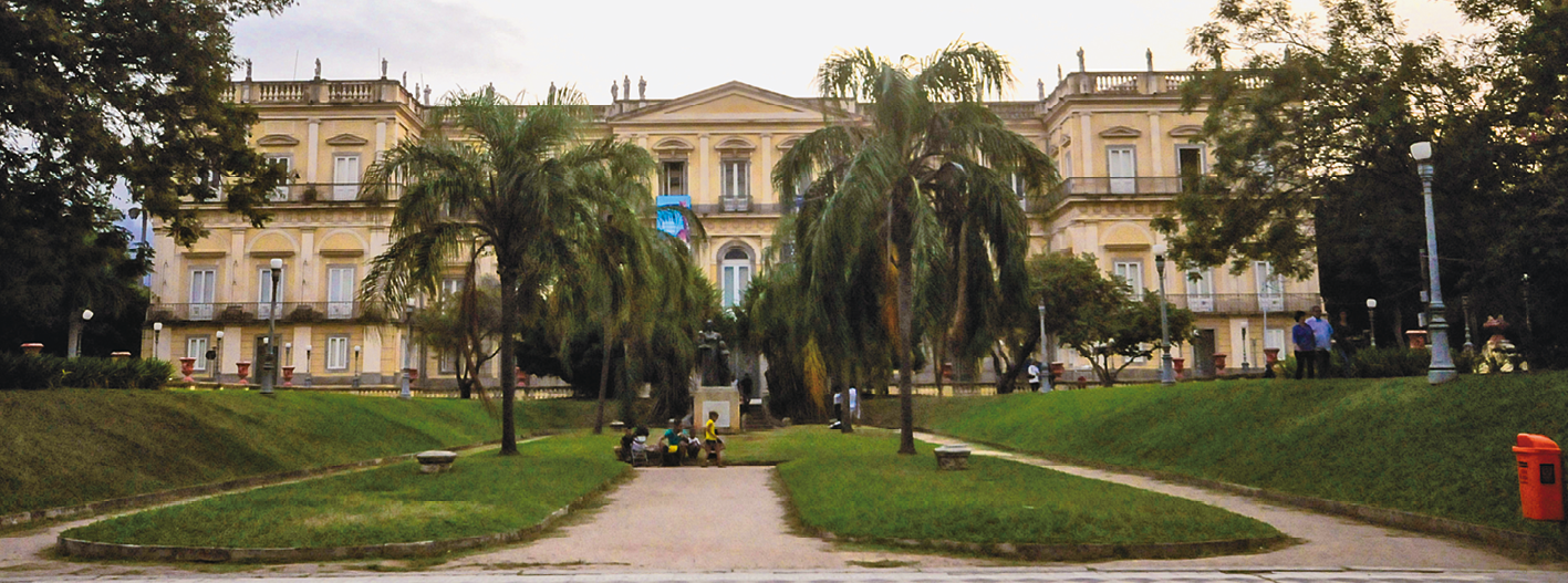 Fotografia. À frente, um amplo espaço com áreas gramadas e muitas árvores. Ao fundo, grande construção com vários andares, grandes portas e janelas, além de uma sacada na parte superior. Há algumas pessoas circulando pelo local.