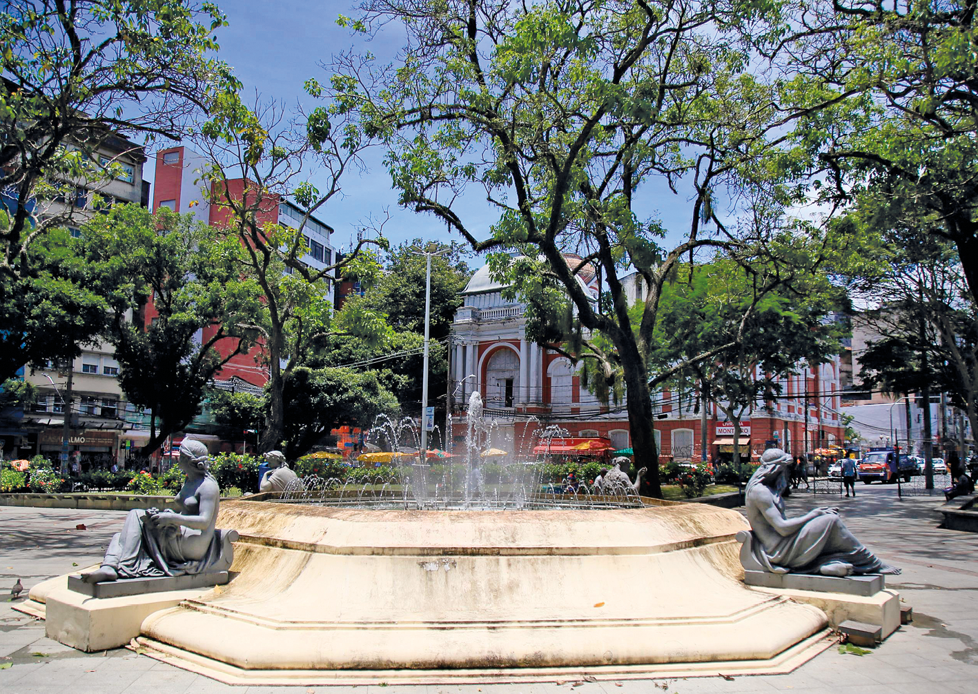 Fotografia. Vista de uma praça com um chafariz e, ao redor, várias estátuas. Há muitas árvores espalhadas pelo lugar e, ao fundo, algumas construções.