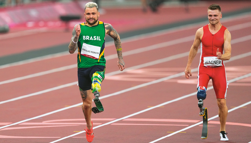 Fotografia de dois homens correndo em uma pista de corrida. Mais à frente homem branco, com cabelos loiros e uniforme verde escrito Brasil. Atrás, homem branco, cabelos castanhos e uniforme vermelho. Ambos usam prótese em uma das pernas.