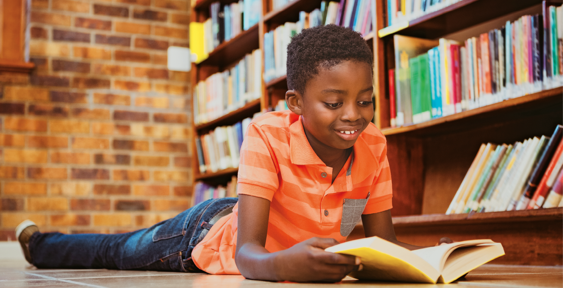 Fotografia. Um garoto negro deitado no chão de bruços, sorrindo e olhando para um livro aberto que está segurando. Ao fundo, prateleiras com livros dispostos lado a lado.