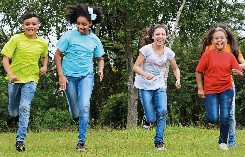 Fotografia. Quatro crianças correndo em uma área ao ar livre, gramada, uma ao lado da outra e todas sorrindo. Da esquerda para a direita: menino branco com camiseta verde, menina negra com camiseta azul, menina branca com camiseta branca e menino negro com camiseta vermelha. Ao fundo, árvores.