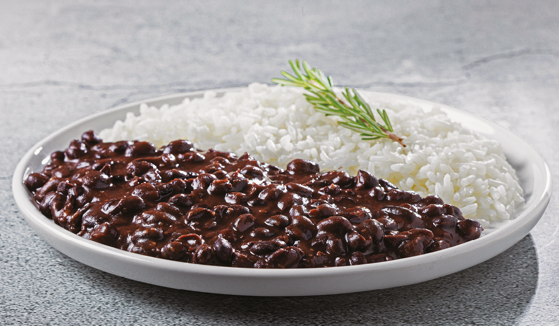 Fotografia. Um prato com feijão preto de um lado e arroz branco do outro lado. Sobre o arroz, um ramo de alecrim.