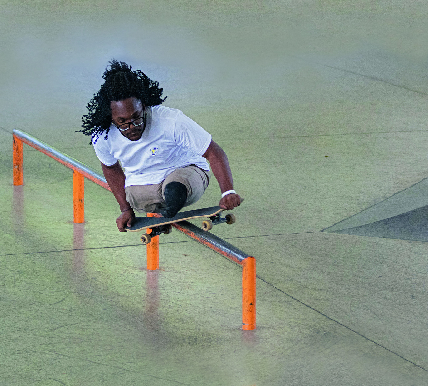 Fotografia. Um rapaz negro de cabelos pretos encaracolados sobre um skate, realizando uma manobra sobre uma barra fina. Ele não possui as pernas e segura o skate com as duas mãos.
