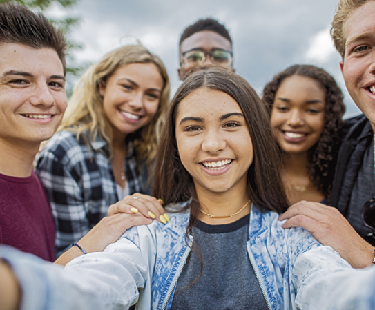 Fotografia. Uma adolescente branca com os braços estendidos para a frente. Ao redor cinco adolescentes de várias etnias.