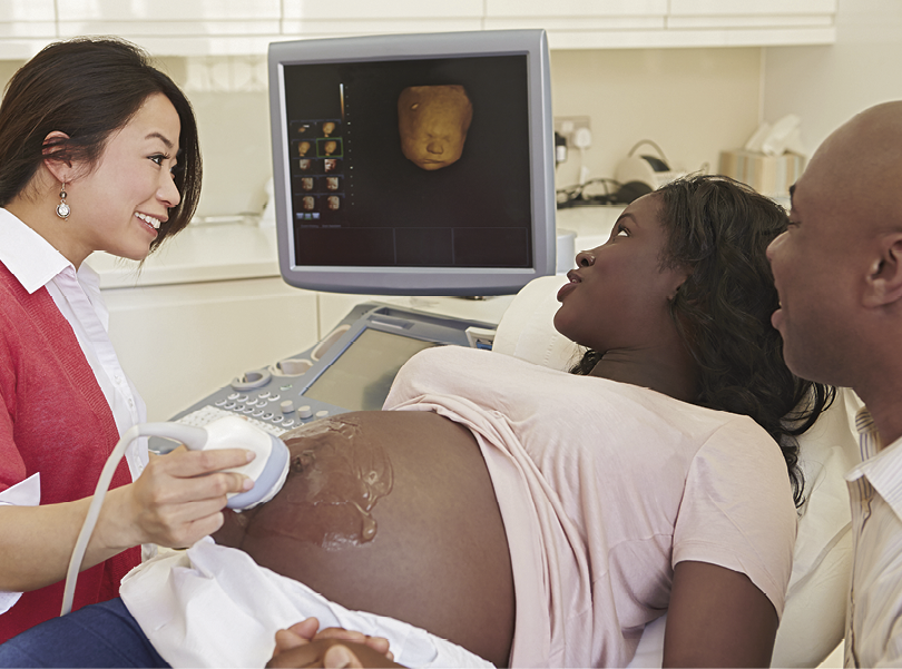 Fotografia. À esquerda, uma mulher branca com cabelos pretos lisos sentada. Com a mão direita ela segura um aparelho que encosta na barriga de uma mulher negra gestante com cabelos pretos encaracolados que está ao seu lado e deitada. Ao lado da mulher gestante há um homem negro sem cabelos. Todos olham na direção de um monitor onde é projetado o rosto de um feto.