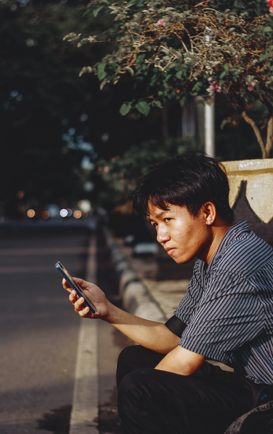 Fotografia. Garoto asiático de cabelos pretos lisos sentado em uma calçada, segurando um celular com a mão direita. Ele olha para a esquerda. Ao fundo, a rua está escura e há vegetação.