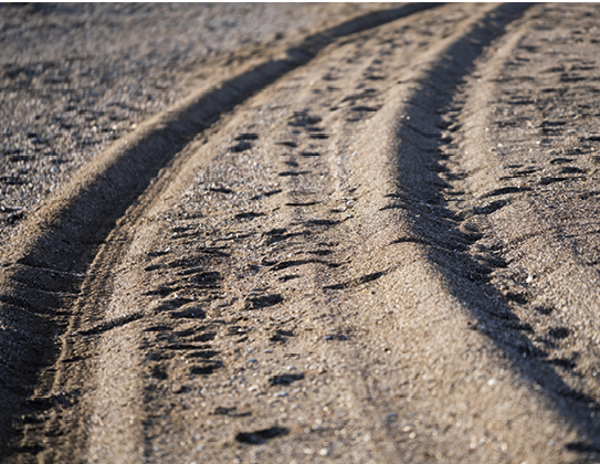 Fotografia. Marcas de dois pneus sobre areia, indicando que um carro passou por ali.