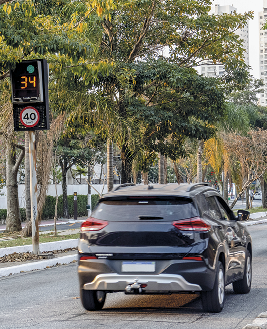 Fotografia. Traseira de um carro preto em uma rua arborizada. À esquerda, uma placa sinalizando 40 quilômetros por hora e, acima o radar marcando 34.