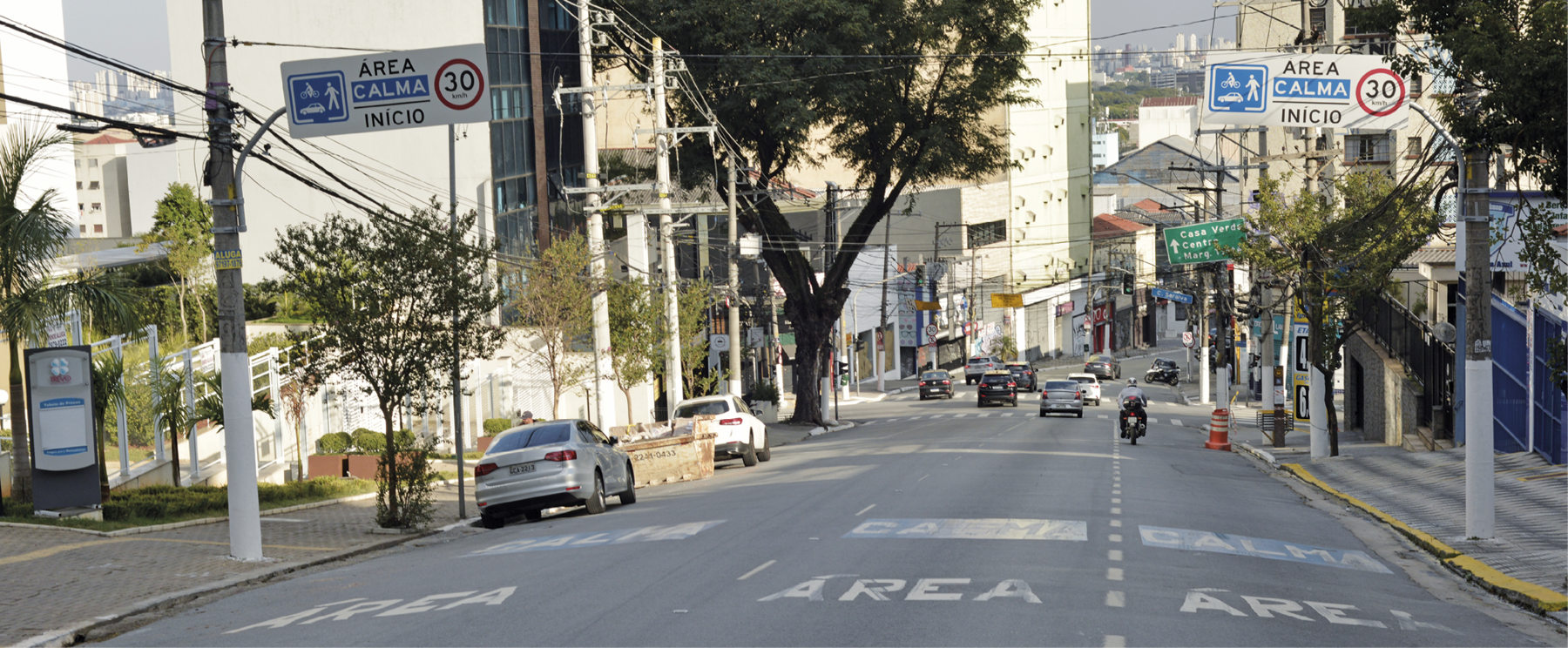 Fotografia de uma rua ampla com alguns carros estacionados e alguns carros andando mais à frente. Há duas placas nas laterais da rua, com a mensagem área calma, início, 30 quilômetros por hora.