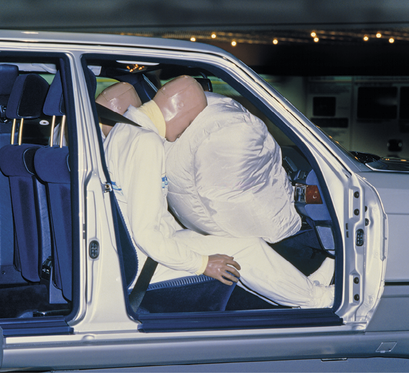 Fotografia que mostra a parte frontal de um carro onde estão bonecos com proporções humanas sentados nos bancos, usando cinto de segurança e, à frente deles, o airbag acionado, um grande saco totalmente inflado.