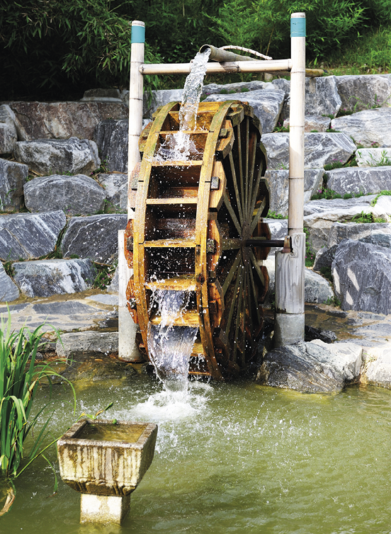 Fotografia de uma roda de madeira com placas na superfície. Cai água na parte superior dela que a faz girar e, na parte inferior, trecho de água. Ao fundo, muro de pedra.
