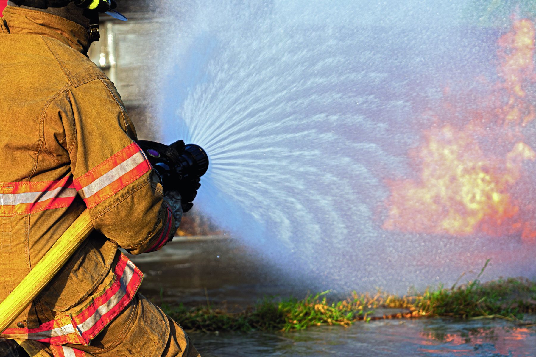 Fotografia. Uma pessoa de costas, usando roupas de proteção, segura uma mangueira que solta um amplo jato de água na direção de um incêndio.