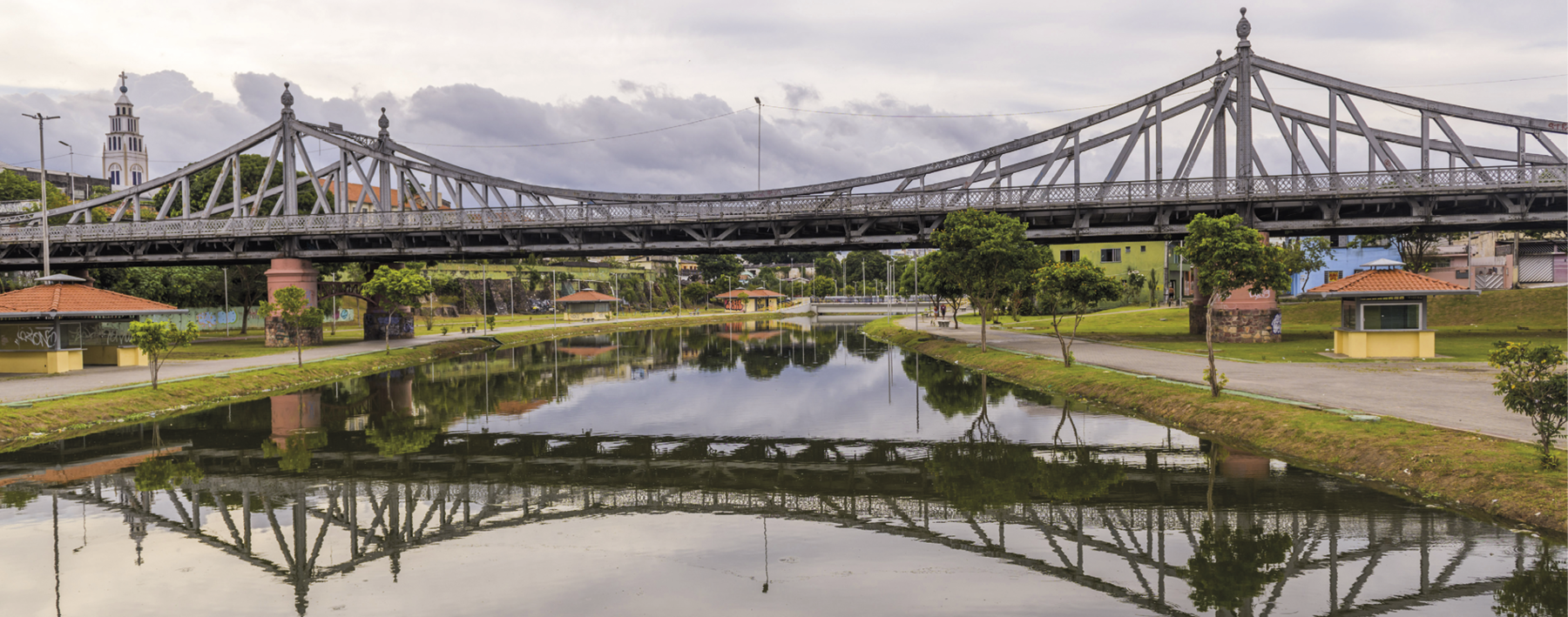 Fotografia. Vista de um lugar por onde passa um rio. Acima, uma ponte com colunas e barras de ferro ao redor, que está refletida na água. Ao redor, árvores e casas