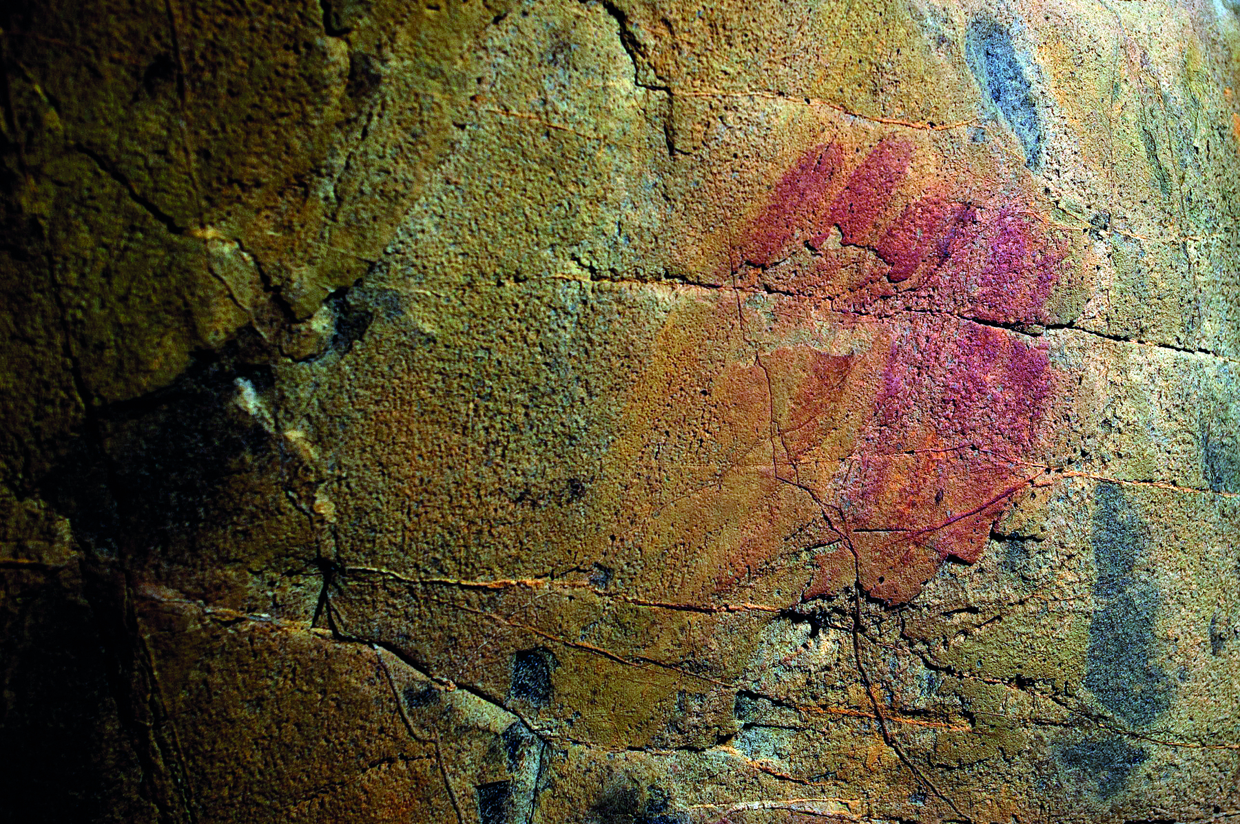 Fotografia. Em uma parede rochosa com algumas ranhuras, manchas avermelhadas e manchas azuladas.