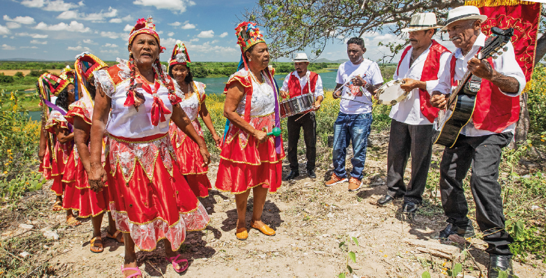 Fotografia. Várias pessoas negras em ambiente aberto com terra e vegetação diversa. À direita, homens vestindo coletes vermelhos e tocando os instrumentos violão, tambor, triângulo e pandeiro. À esquerda, mulheres usando blusa branca, saia vermelha, calçados e chapéus com fitas coloridas. Ao fundo, céu azul com algumas nuvens brancas, vegetação e trecho com água.