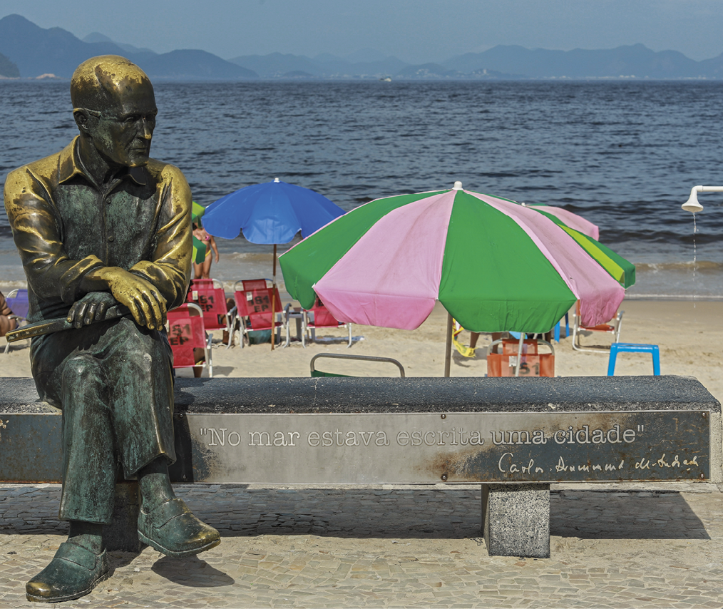 Fotografia. Estátua de homem sentado em um banco com as pernas cruzadas e as mãos sobre a coxa, olhando para o lado. Ao fundo, areia, guarda-sóis e mar. A estátua é bronzeada com manchas azuladas.