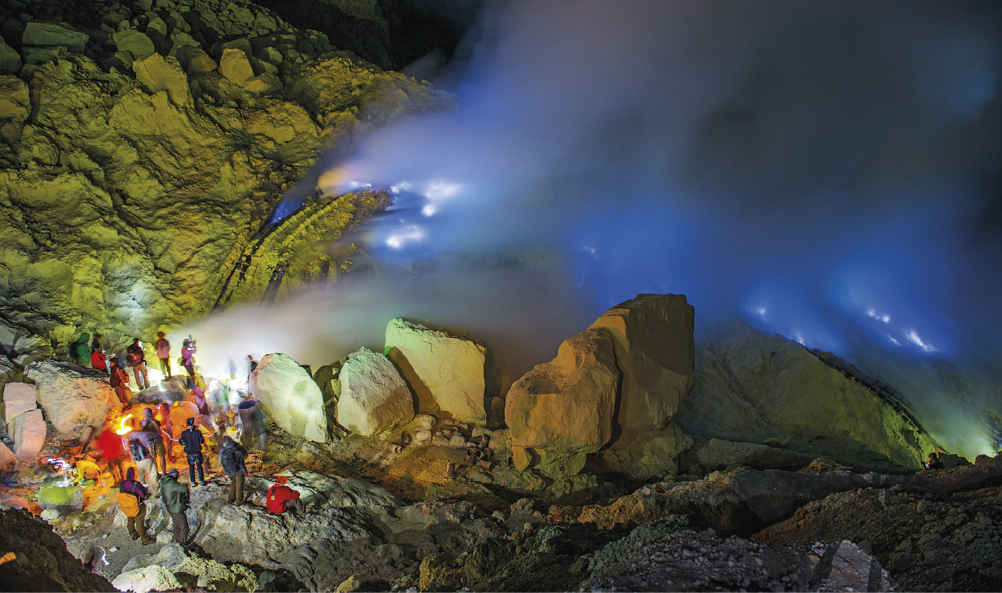 Fotografia que mostra algumas pessoas próximas à boca de um vulcão, com pedras ao redor. No centro do vulcão, fumaça azul e branca.