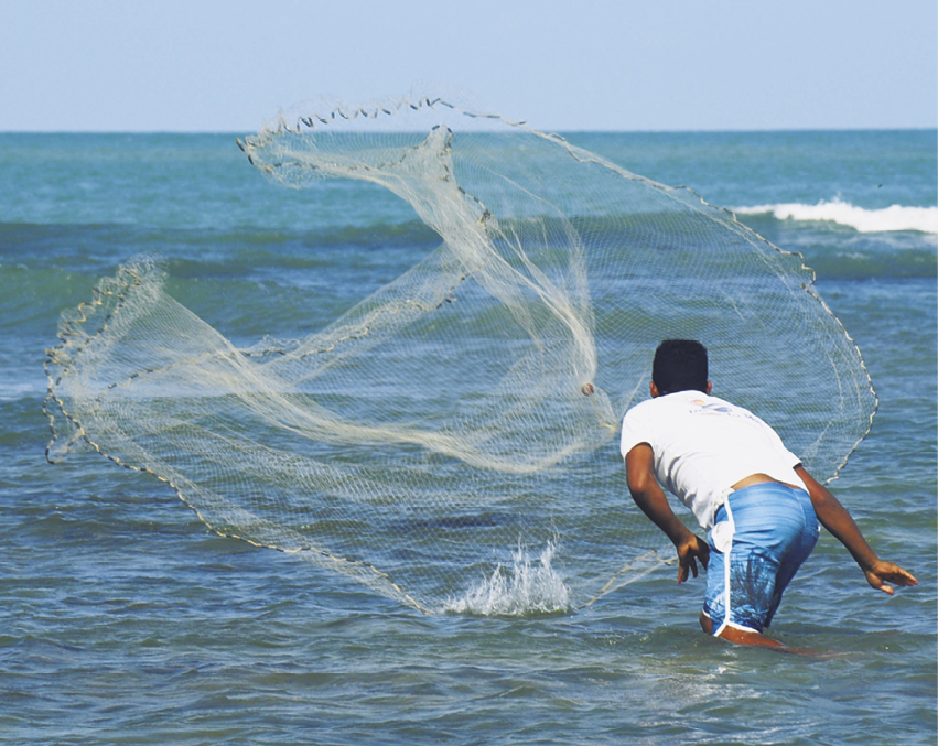 Fotografia. Um homem em meio ao mar, lançando uma rede de pesca. A rede está aberta no ar, prestes a cair na água.