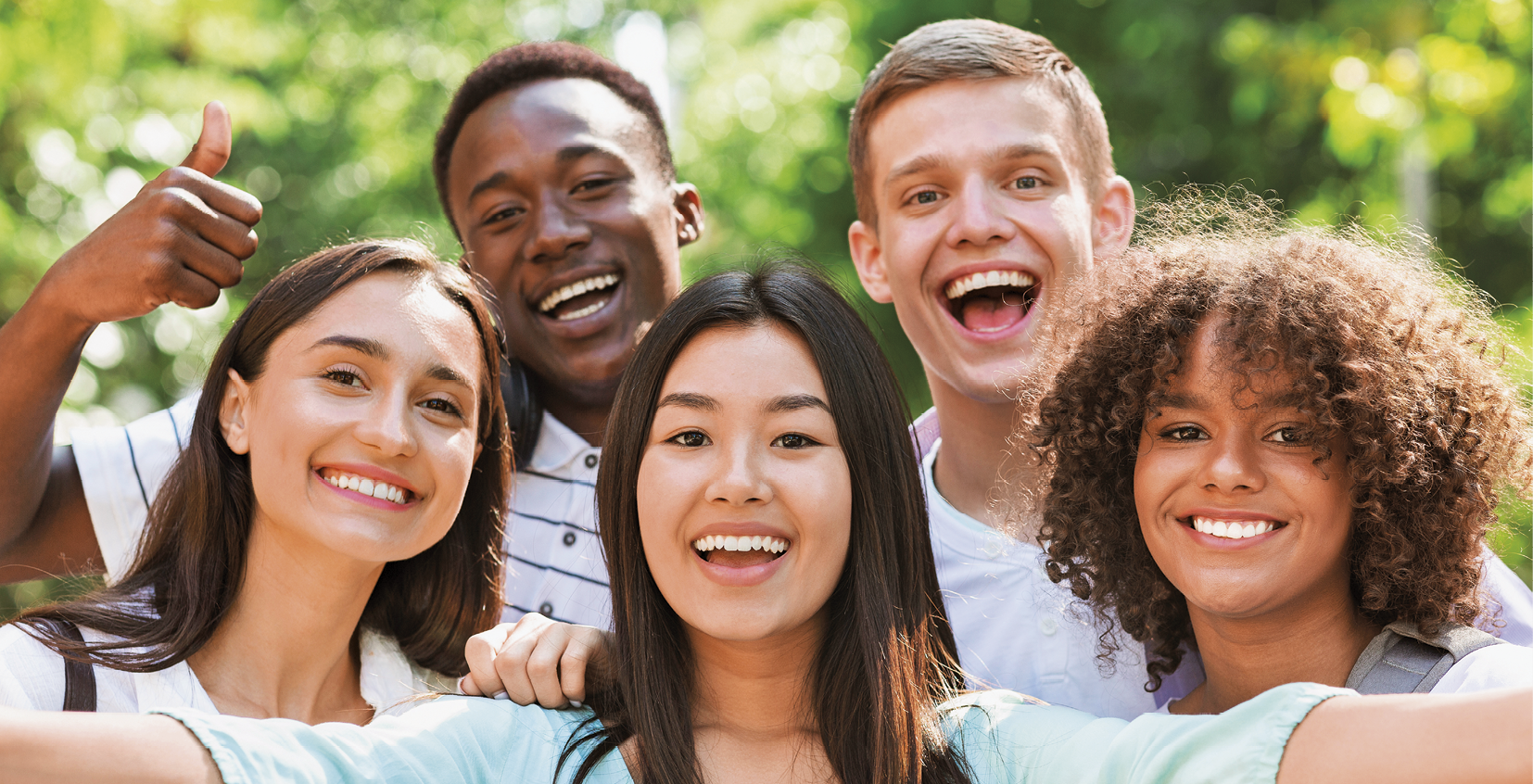 Fotografia que mostra cinco pessoas sorridentes vistas do ombro para cima. À frente, uma mulher branca com cabelo escuro, uma mulher asiática com os braços abertos e uma mulher negra. Atrás, um homem negro com uma mão levantada com polegar para cima e um homem branco.