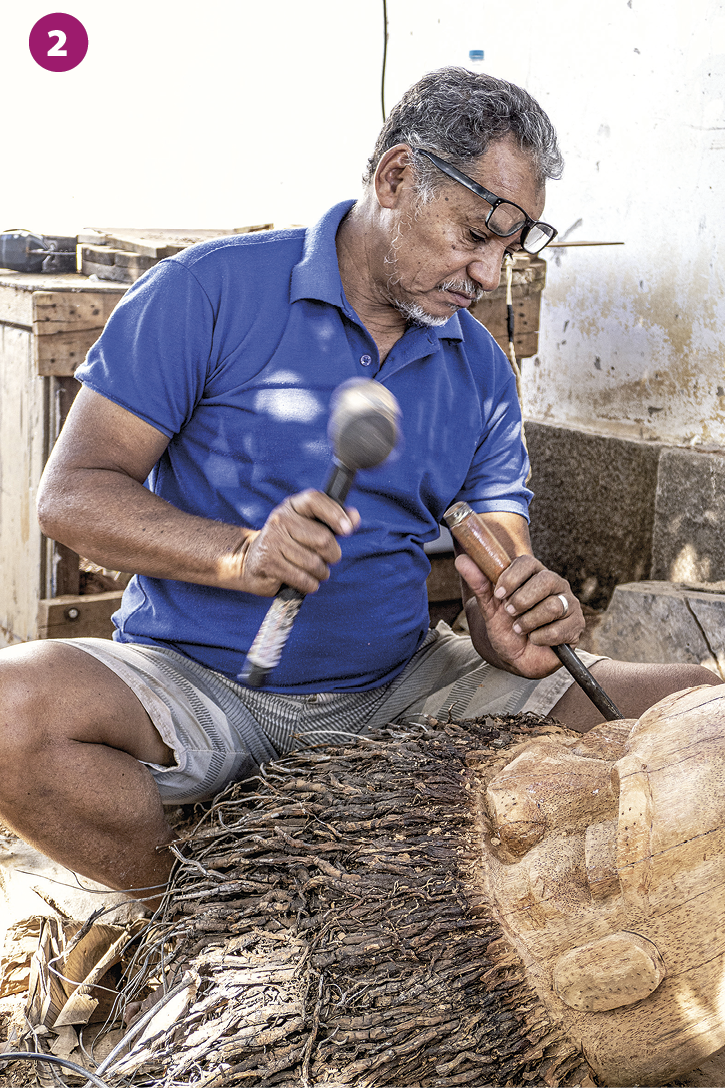 Fotografia 2. Um homem de cabelos grisalhos, usando óculos, camisa azul e shorts, esculpe um tronco de madeira com um martelo e um cinzel, instrumento de metal utilizado especificamente para entalhar.