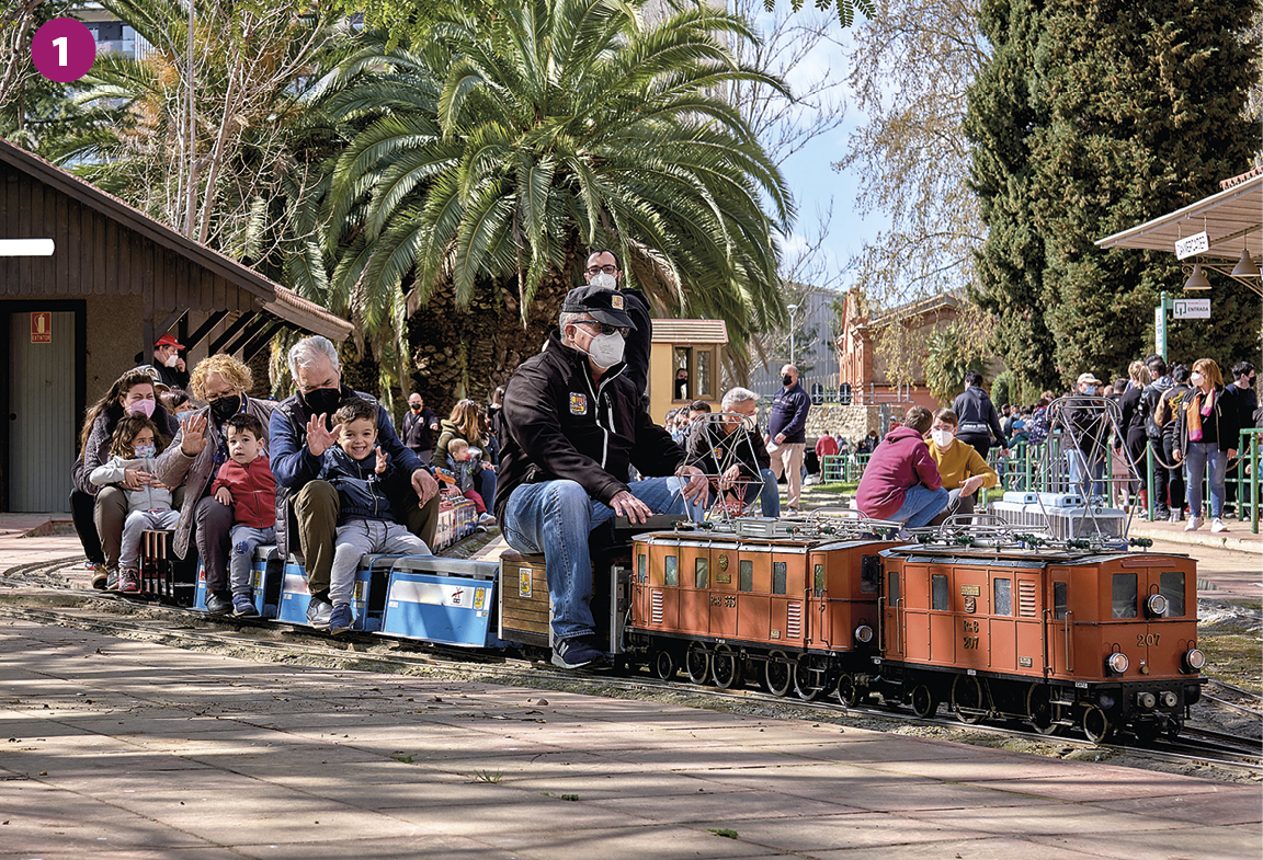 Fotografia 1. Diversos adultos e crianças agasalhados com casacos estão sentados sobre um trem pequeno de brinquedo, com as cores em tons de vermelho e azul. Eles estão sorrindo e com os braços levantados. Ao fundo há pessoas em pé e sentadas, árvores e pequenas construções.