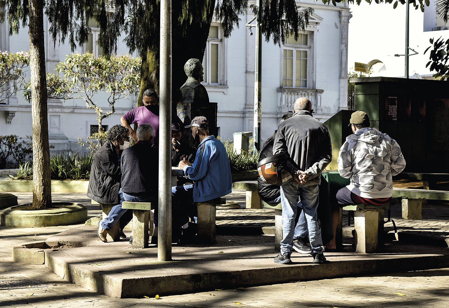 Fotografia. Pessoas reunidas em uma praça. Elas estão sentadas ao entorno de duas mesas, e duas pessoas estão em pé observando o restante do grupo. Ao fundo há árvores e uma grande construção branca.