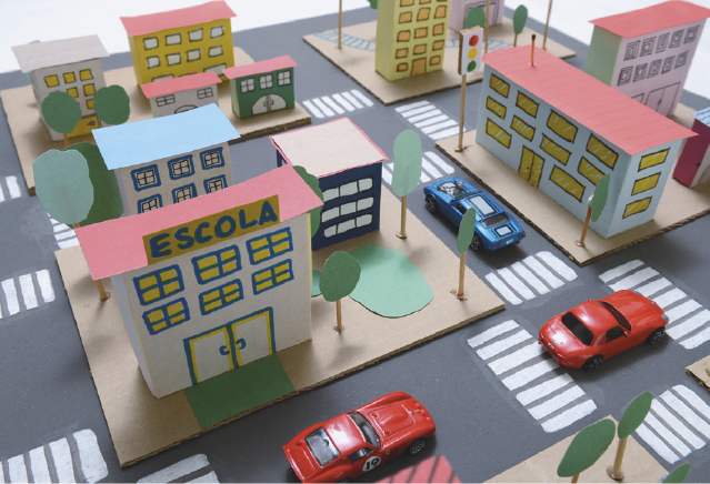 Maquete. Representação de uma maquete de prédios com janelas e telhados. No centro há um prédio com a inscrição ESCOLA em uma placa. Ao redor, ruas com faixas de pedestre, carros e árvores nas calçadas.