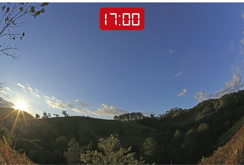 Fotografia. Vista de uma uma paisagem com declives, gramado verde e algumas árvores na parte inferior. Na parte superior, o céu está azul e o sol está posicionado ao lado esquerdo, quase oculto pelo declive e morros presentes na paisagem. A vegetação está em sua maioria sombreada. No topo da imagem, um visor de relógio digital indica o horário: 17 horas.
