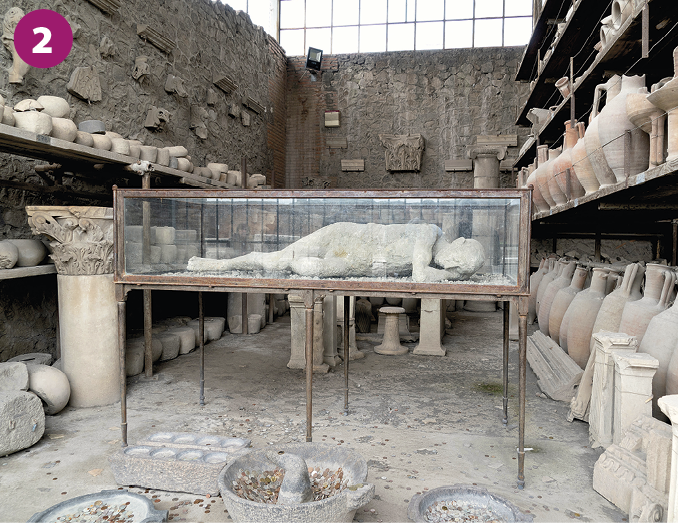 Fotografia 2. Um corpo humano em tons de cinza dentro de uma caixa de vidro. Ao redor há diversos vasos e objetos de cerâmica em tons amarronzados.