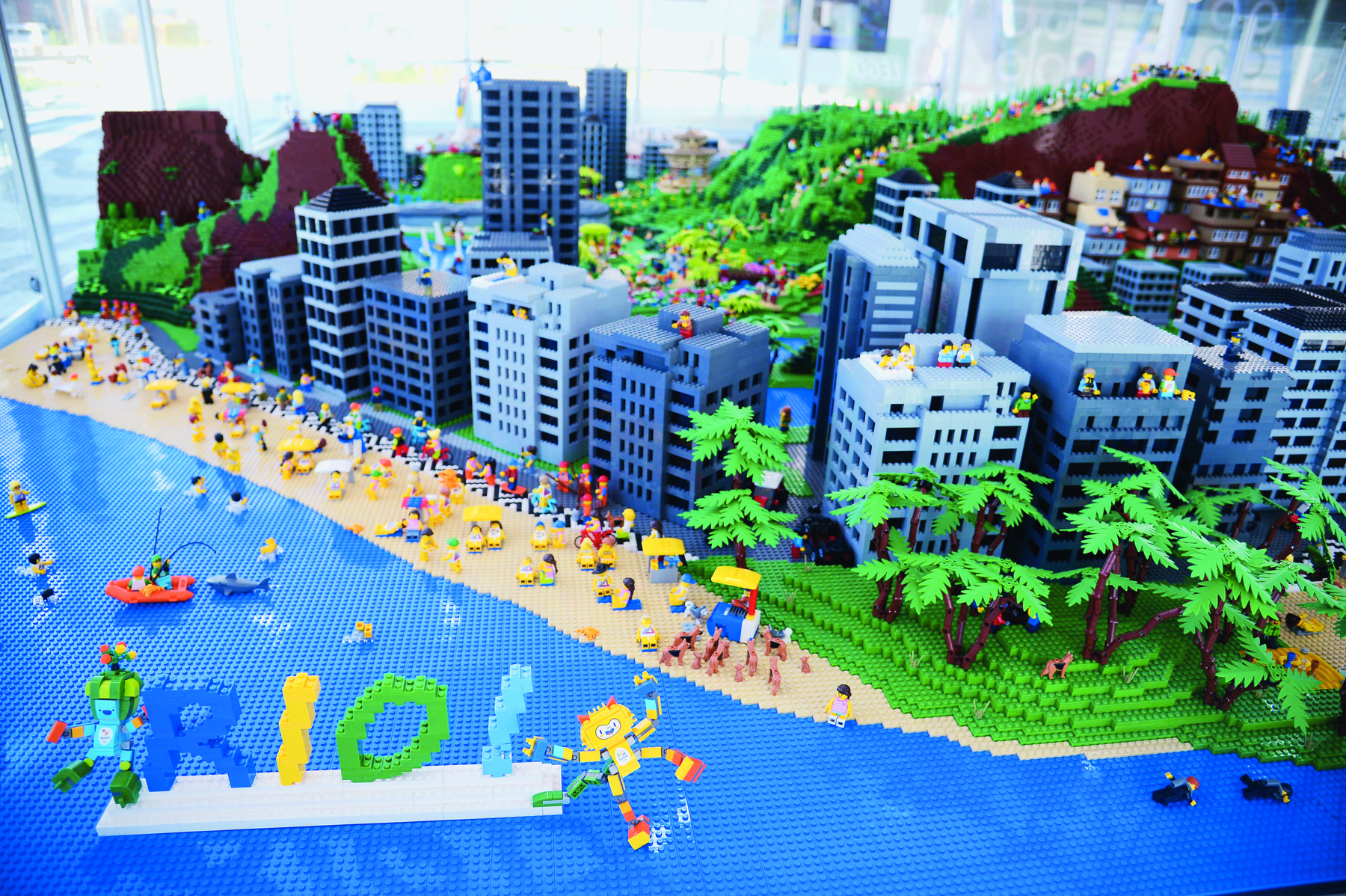 Maquete. A imagem mostra uma maquete, construída com peças de montar, que representa a orla da praia da cidade do Rio de Janeiro, ou seja, com presença de mar, praia e prédios. Ao fundo, morros com construções na encosta.
